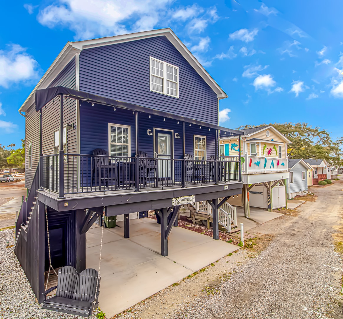 The raised beach house is depicted with a dark navy blue exterior, featuring a wide deck with railing and multiple outdoor seating options. The area below offers paved parking spaces, complemented by chairs on the deck and neighboring properties visible in the background under a clear blue sky.