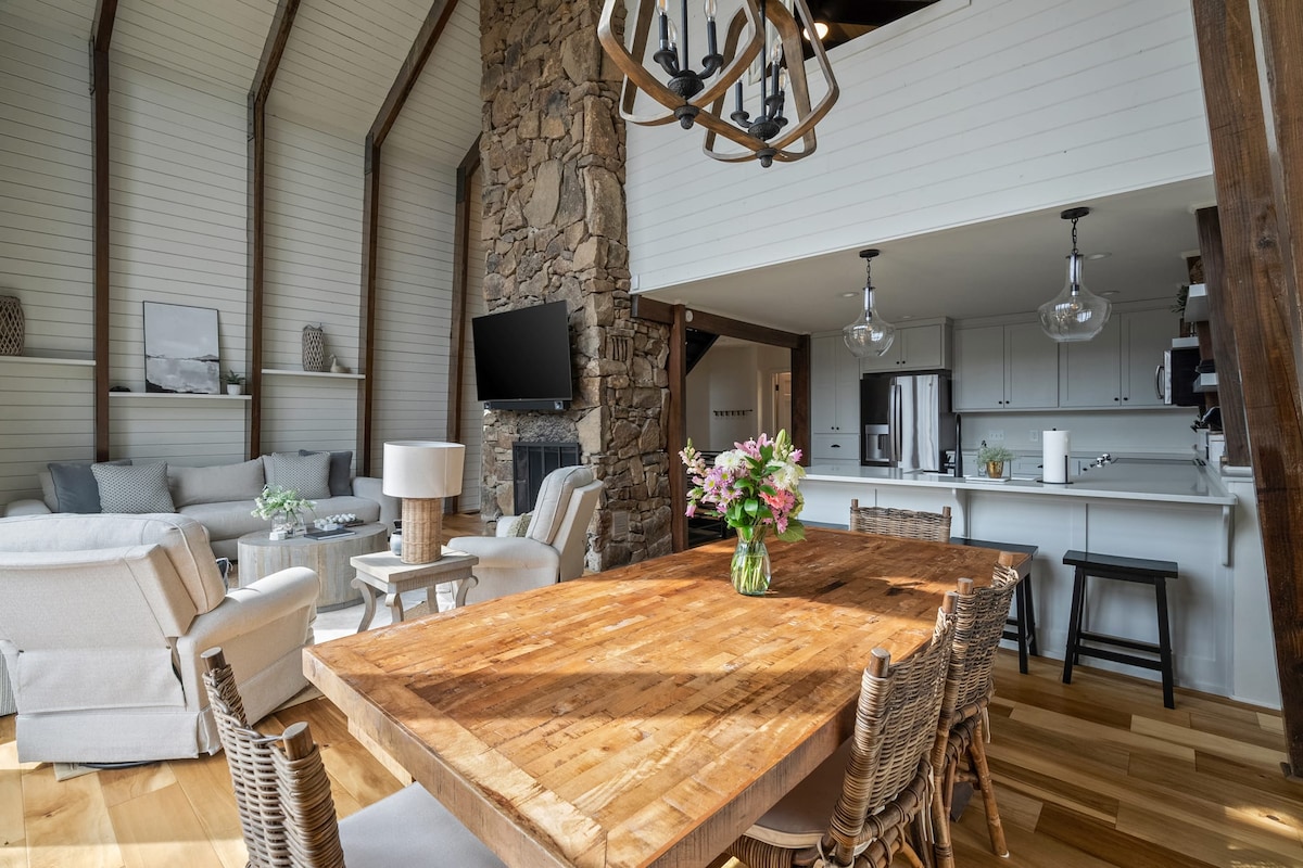 A spacious living and dining area features a large wooden dining table surrounded by woven chairs. A comfortable seating arrangement is positioned nearby, accompanied by a flat-screen television mounted on a stone wall. Natural light streams in through high windows, enhancing the open atmosphere.