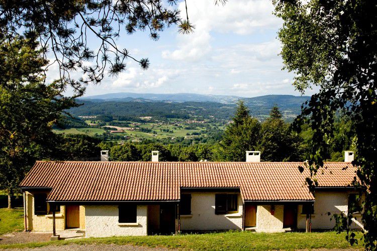 A hillside accommodation is visible, featuring a row of single-story units with sloped roofs. Large windows allow for natural light, and the surrounding landscape of rolling hills and greenery is prominent, providing a serene backdrop under a partly cloudy sky.