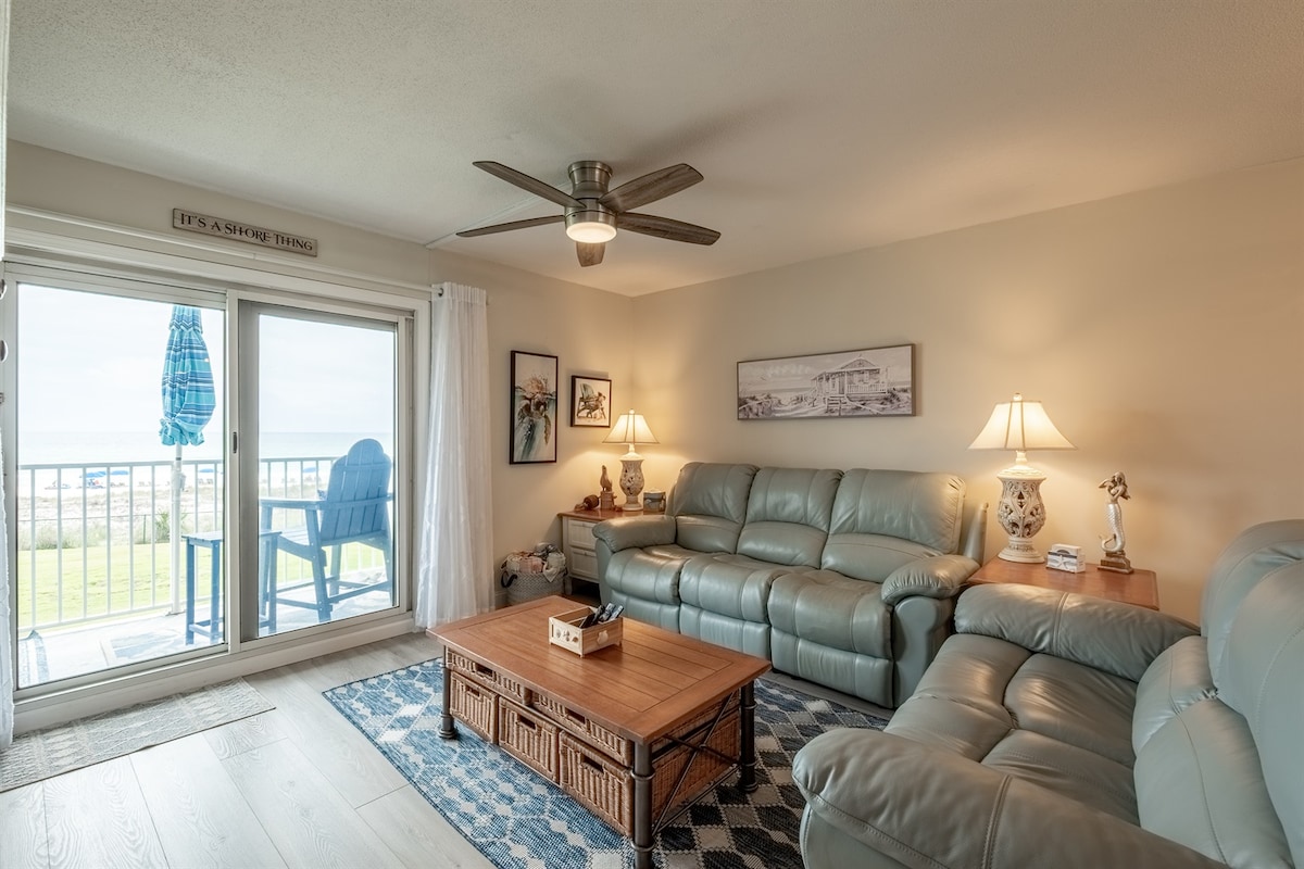 A comfortable living area features two light blue sofas arranged around a wooden coffee table, creating a cozy seating space. A ceiling fan is present, and natural light enters through large sliding glass doors, which lead to a balcony with a view of the outdoors.