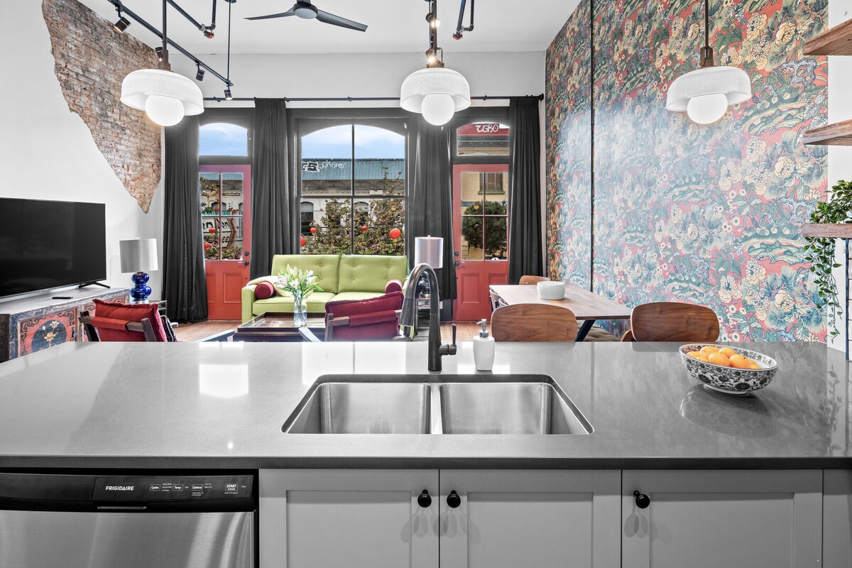A modern kitchen features sleek countertops and a double sink, with stylish pendant lights overhead. In the background, a vibrant living area is visible, adorned with colorful furnishings and large windows that fill the space with natural light.