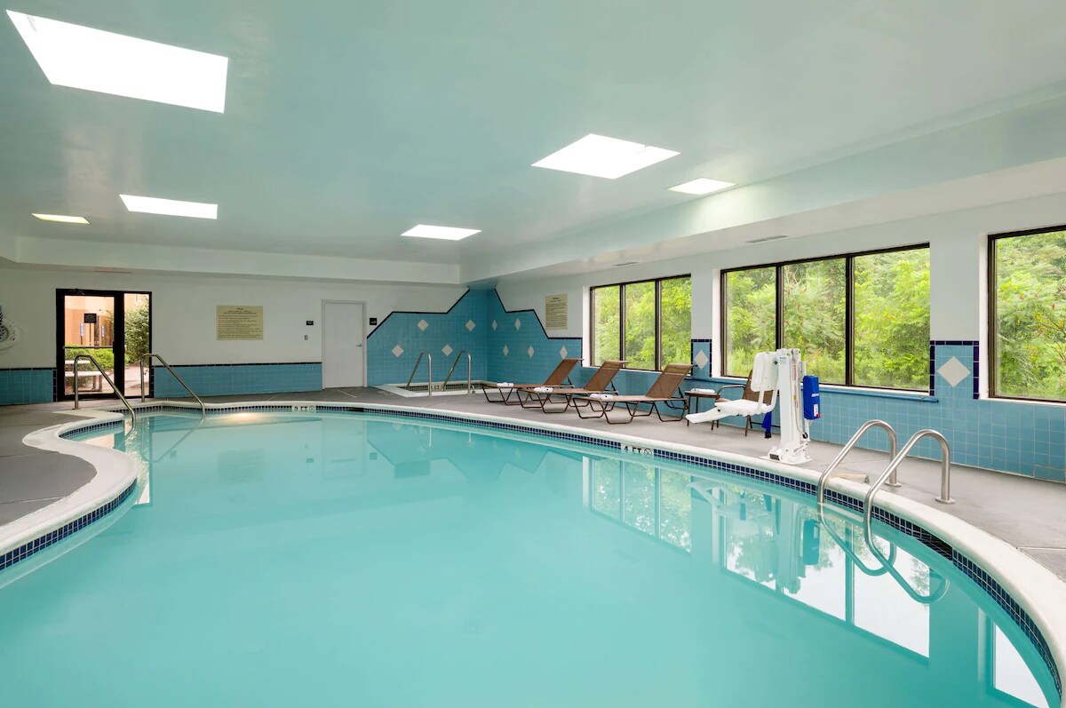 An indoor pool is illuminated by natural light from large windows and skylights, revealing a blue-tiled area surrounded by several lounge chairs. The pool is inviting with a clear, turquoise surface, while a safety railing is positioned at the shallow end.