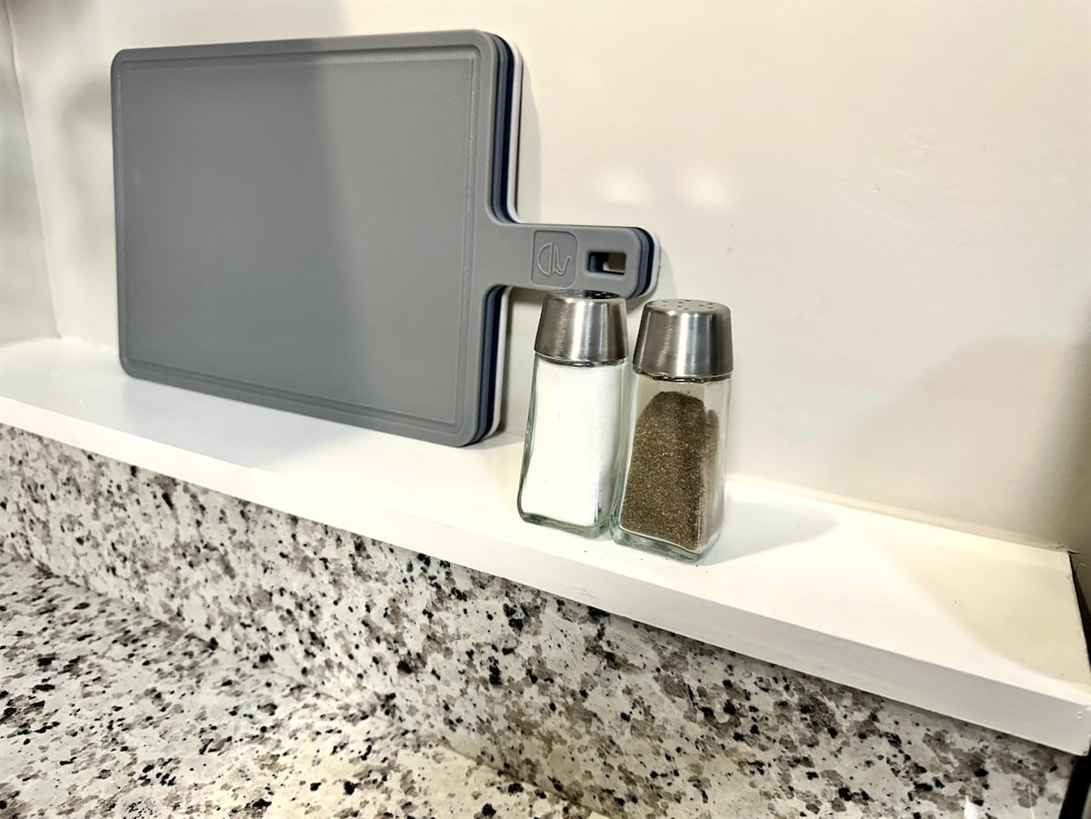 A kitchen countertop features a grey cutting board alongside salt and pepper shakers. The shakers are glass with metal tops, resting on a white shelf that contrasts with the speckled granite surface below.