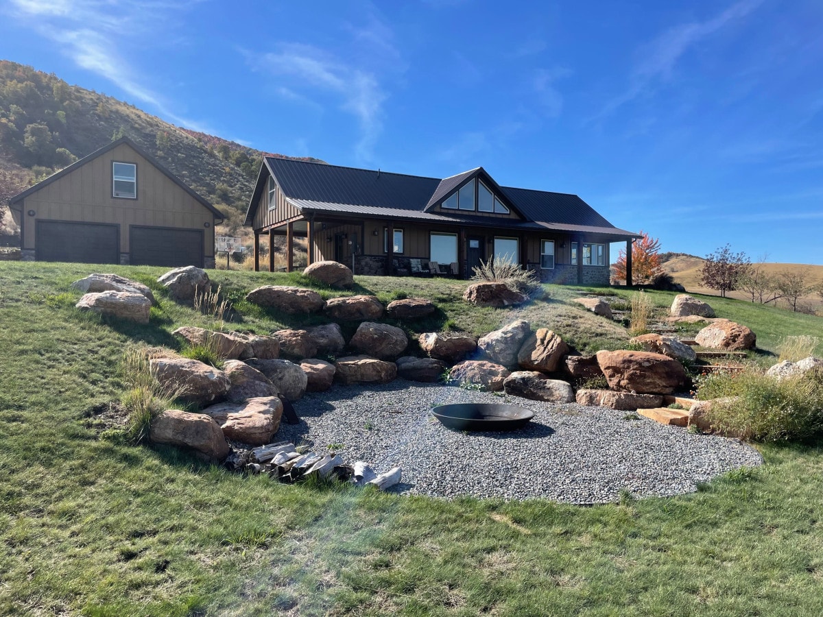 A modern home is set against a hillside, featuring large windows and a dark roof. Surrounding the house are landscaped areas with large boulders and pebbles, leading to a fire pit placed on gravel. Clear skies and natural greenery enhance the rural setting.
