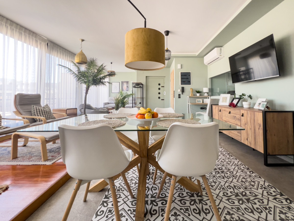 A dining area features a glass table supported by a wooden base, surrounded by white chairs. A bowl of colorful fruit sits at the center. Natural light fills the space, highlighting a large decorative rug and modern furniture, including a stylish lamp overhead.