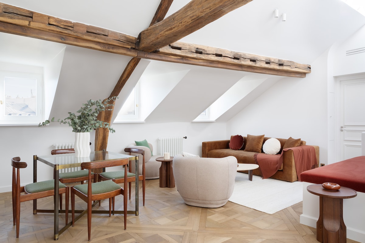 An airy living space is highlighted by exposed wooden beams and a combination of plush seating options, including a cozy sofa and armchairs. A dining table with green accents sits adjacent to the lounge area, while light pours in through windows, enhancing the elegant design.