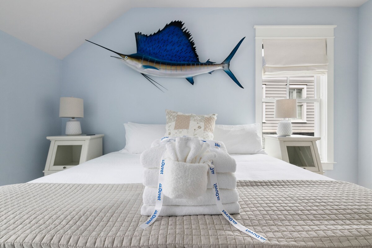 A spacious bedroom is highlighted by a large blue sailfish mounted on the wall. Soft white linens cover the bed, accompanied by neatly stacked plush towels on a gray quilt at the foot. Natural light flows in from the window, enhancing the serene atmosphere.