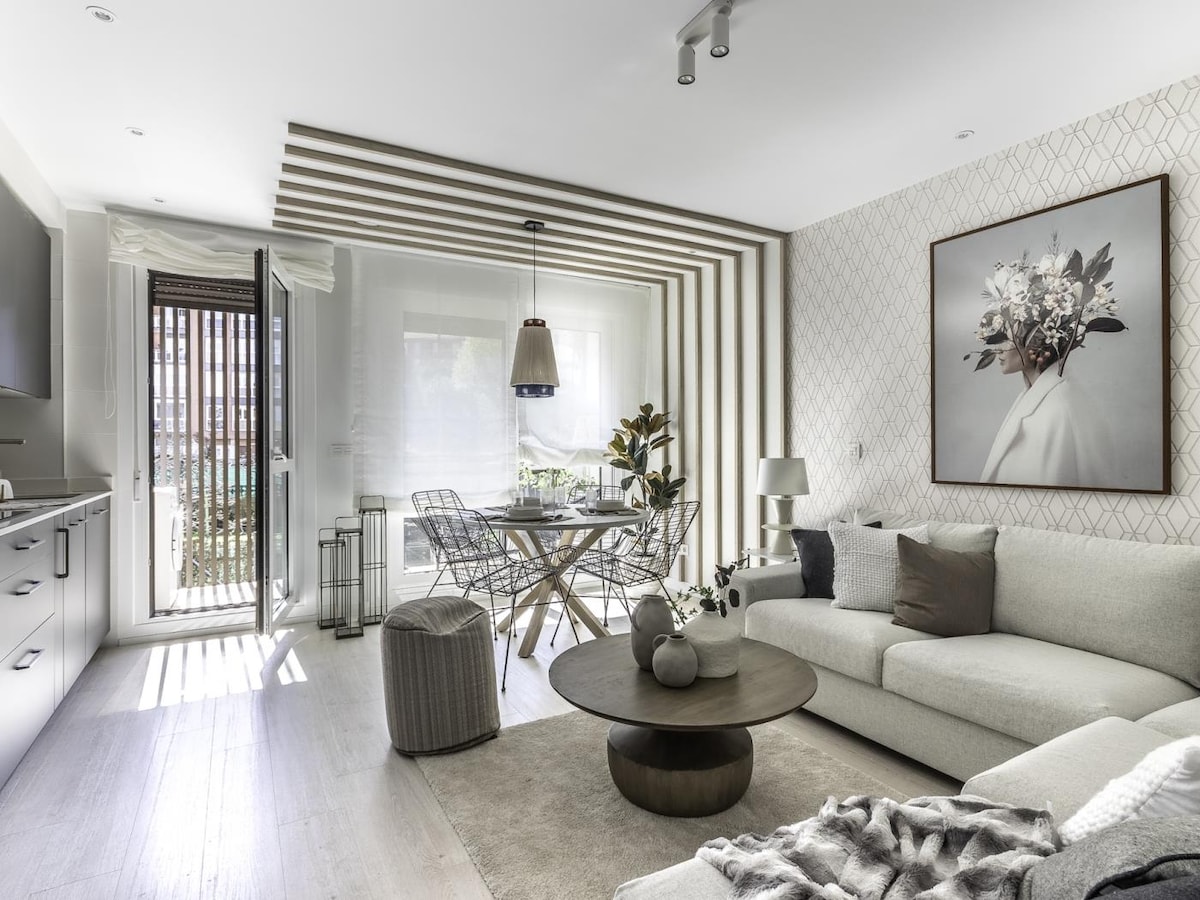 A modern living space is highlighted by a light-colored sofa and a round coffee table. A dining area with metal chairs is visible near large glass doors, allowing natural light to enter. Decorative plants and a framed artwork add a touch of elegance to the room.