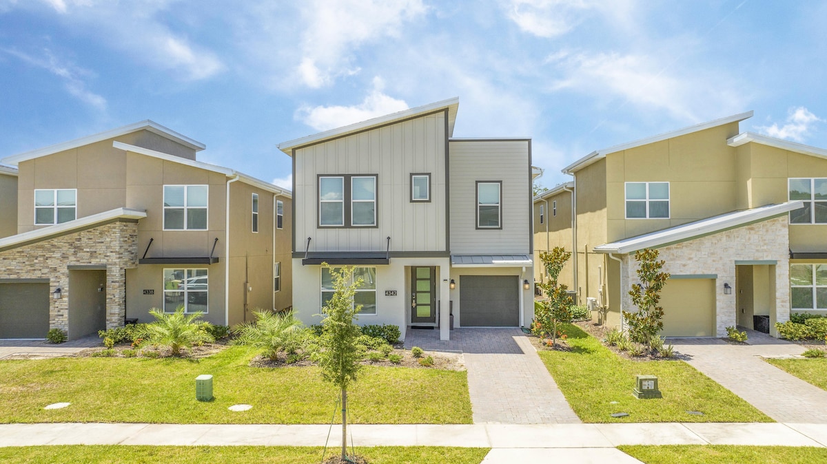 The exterior of a modern two-story home is displayed, showcasing a neutral color palette and contemporary architectural design. Lush greenery frames the property, while a neatly paved pathway leads to the entrance. Adjacent homes feature varied designs, contributing to a vibrant neighborhood atmosphere.