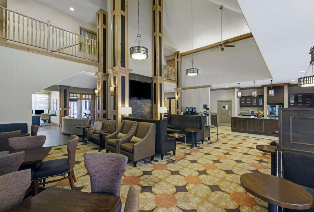 The lobby area features a spacious layout with high ceilings and wood accents. Cozy seating arrangements, including armchairs and small tables, provide inviting spaces for relaxation. A large flat-screen TV is mounted on the wall, complementing the warm color palette of the carpet and decor.