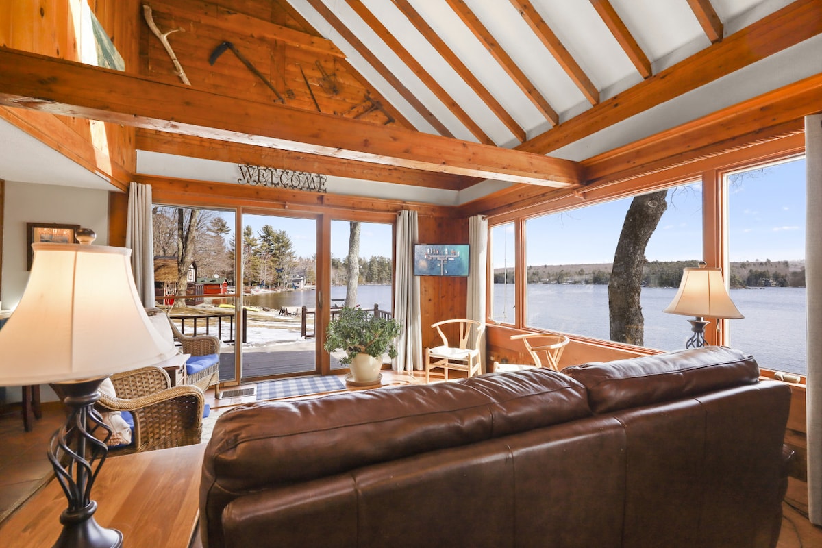 A cozy living area features wooden beams and large windows providing views of the lake. Comfortable seating is arranged around a coffee table, with a warm glow from lamps enhancing the ambiance. The room is designed for relaxation and conversation, seamlessly connecting with the outdoor landscape.