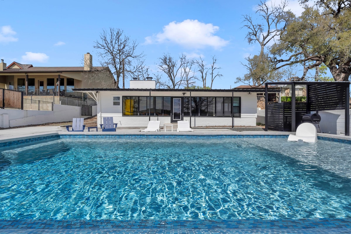 The private pool is surrounded by a spacious patio, featuring lounge chairs positioned for sunbathing. A slide adds a fun element to the pool area, while large glass doors provide a view into the modern interior. Nearby trees offer shade and natural beauty.