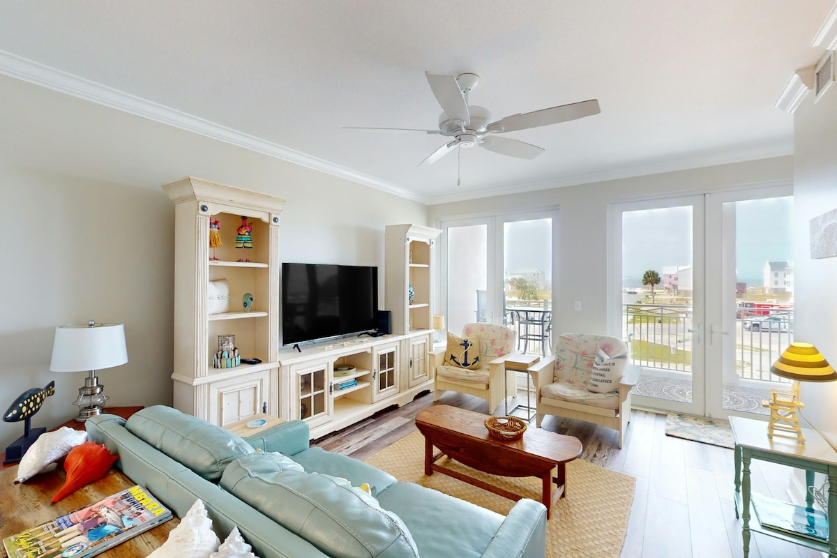 The living area features light-colored walls and a ceiling fan. A comfortable sofa and two armchairs are arranged around a wooden coffee table. A television is mounted on an entertainment unit, with sliding glass doors providing access to a balcony and views of the outdoors.
