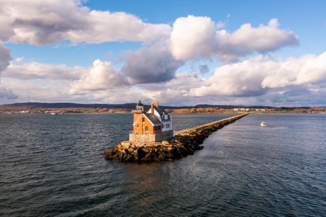 A charming lighthouse stands at the end of a rocky jetty, surrounded by water. The structure features a bright orange exterior with a white roof, contrasted against a backdrop of soft blue skies and fluffy white clouds. Waves gently lap against the stones.