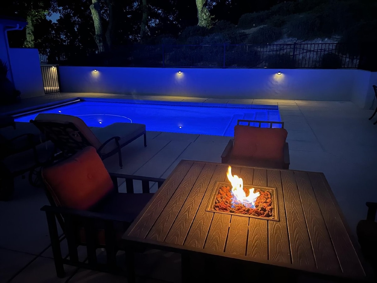 A serene outdoor space is illuminated by soft lighting, highlighting the sparkling pool and a cozy fire pit surrounded by comfortable seating. The flames flicker gently atop the stone-granite table, creating a peaceful ambiance ideal for relaxation at night.