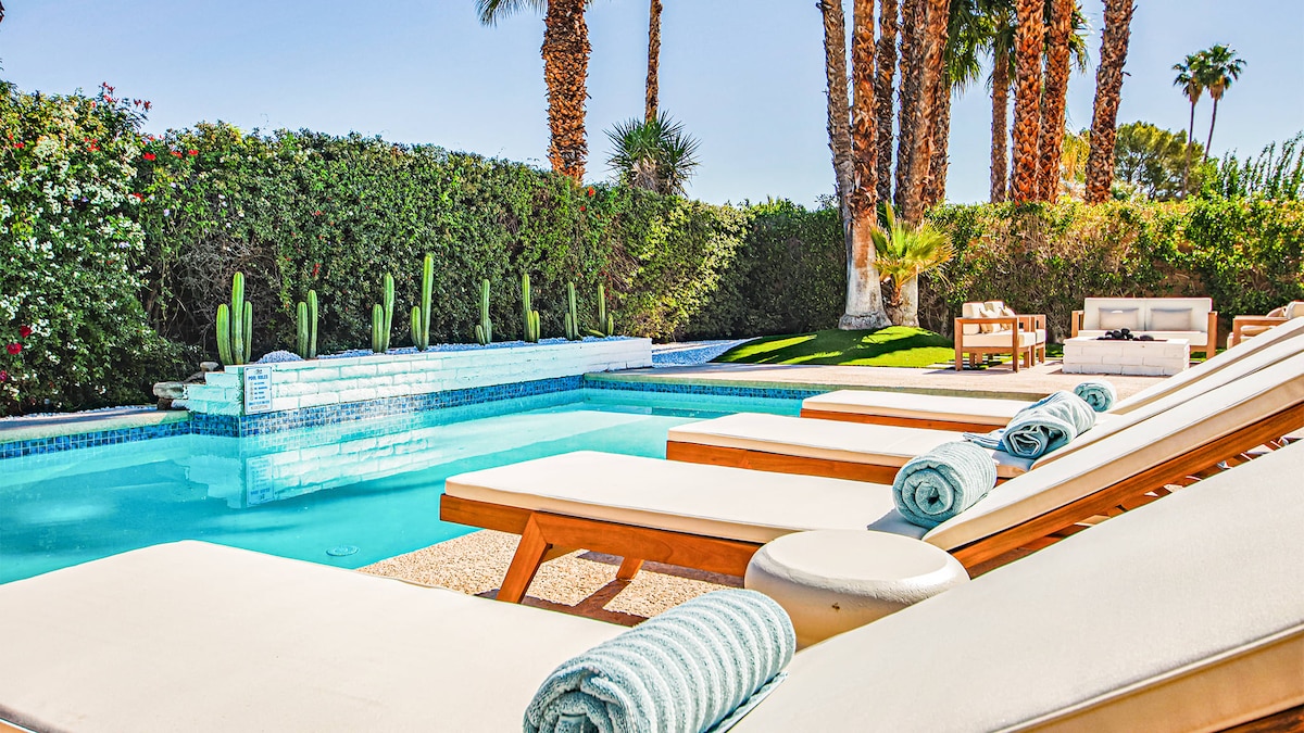 A refreshing swimming pool, surrounded by palm trees, features spacious lounge chairs arranged around it. Each chair is neatly rolled with a towel, inviting relaxation. Lush greenery lines the background, enhancing the inviting outdoor space.