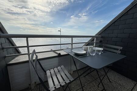 A small terrace features a black table with two folding chairs and an unobstructed view of the sky and sea. Two plates and glasses are neatly arranged on the table, emphasizing the space's inviting atmosphere for outdoor dining.