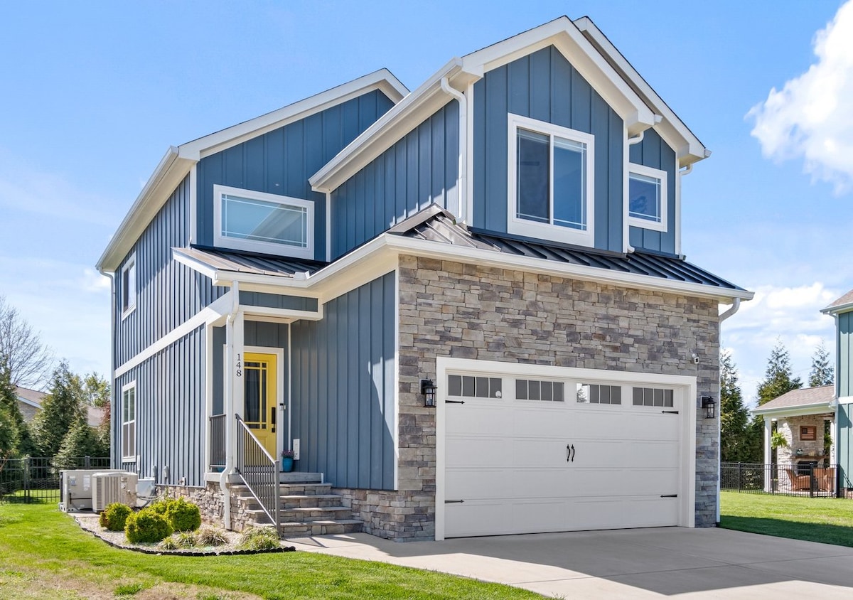 A modern two-story cottage showcases a blend of blue siding and stone accents. Large windows provide natural light, while the spacious driveway leads to a double garage. A well-maintained lawn features organized landscaping with small shrubs and a concrete pathway to the entrance.
