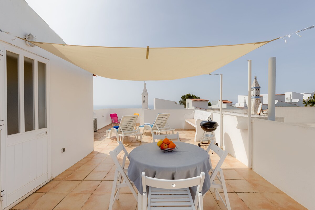A spacious rooftop terrace is presented, featuring a dining table with a grey tablecloth and white chairs. Sun loungers are positioned under a shade sail, with a view of the ocean visible in the background. A barbecue grill is also located on the terrace.