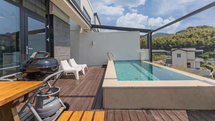 An outdoor terrace features a private plunge pool with clear blue water. A set of light-colored loungers is positioned beside the pool. A charcoal grill is also visible, situated on a wooden deck that offers ample space for relaxation.