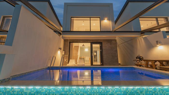 A modern two-story villa is presented with a serene swimming pool featuring tiled edges and soft lighting. Large glass doors provide a view of the interior, where spacious lounge chairs are visible, creating an inviting outdoor area.