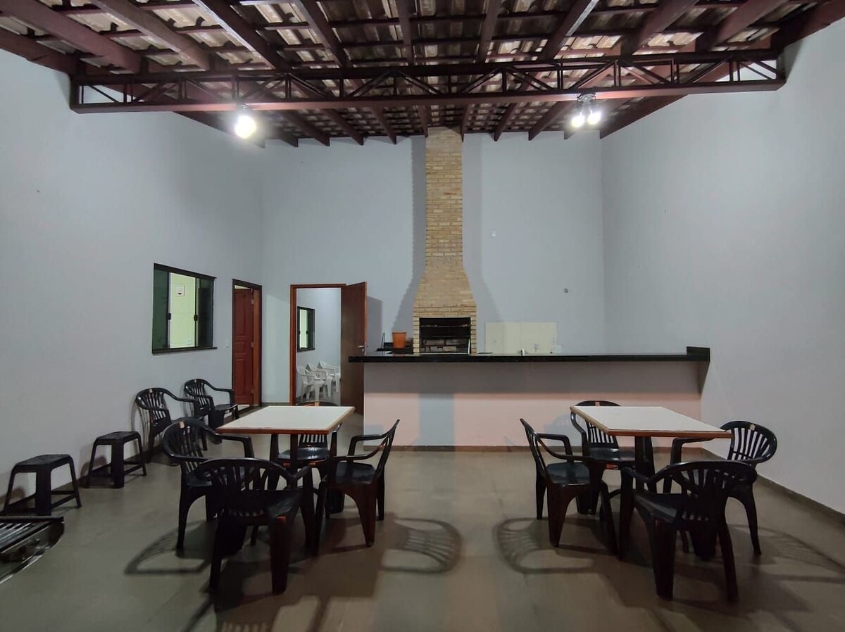 An open gourmet space is presented with two large tables surrounded by black plastic chairs. A stone fireplace is centered along the far wall, adding a focal point to the room. Bright walls and exposed beams create an airy ambiance.