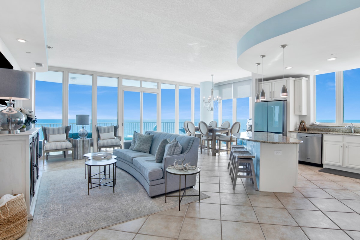 An open-concept living area features large floor-to-ceiling windows that frame panoramic views of the lagoon and Gulf. A spacious sectional sofa and dining area are complemented by clean lines and soft, coastal hues. The kitchen, equipped with modern amenities, offers additional seating options.