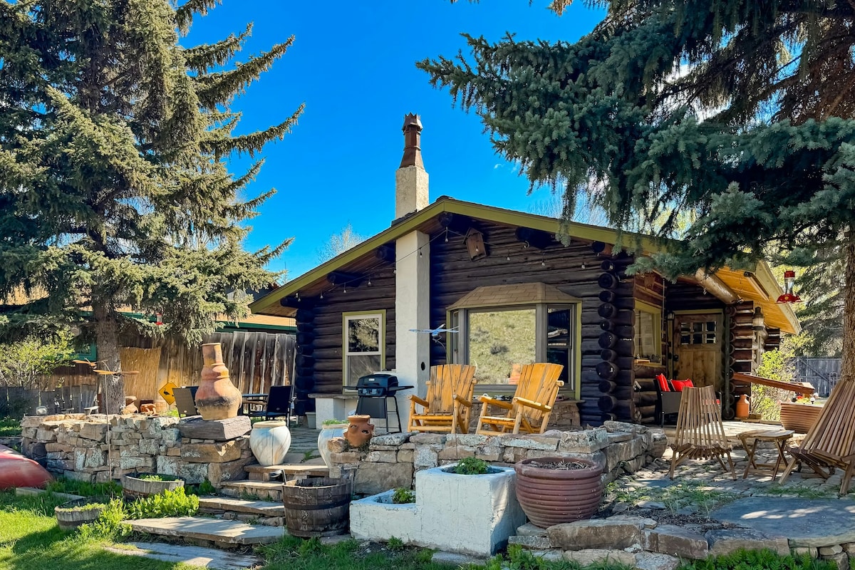 A rustic cabin nestled among tall trees features a welcoming patio area. Multiple wooden chairs surround a stone firepit, with various planters and decorative elements visible. A grill is positioned nearby, while the cabin's log construction and large windows reflect the outdoor landscape.