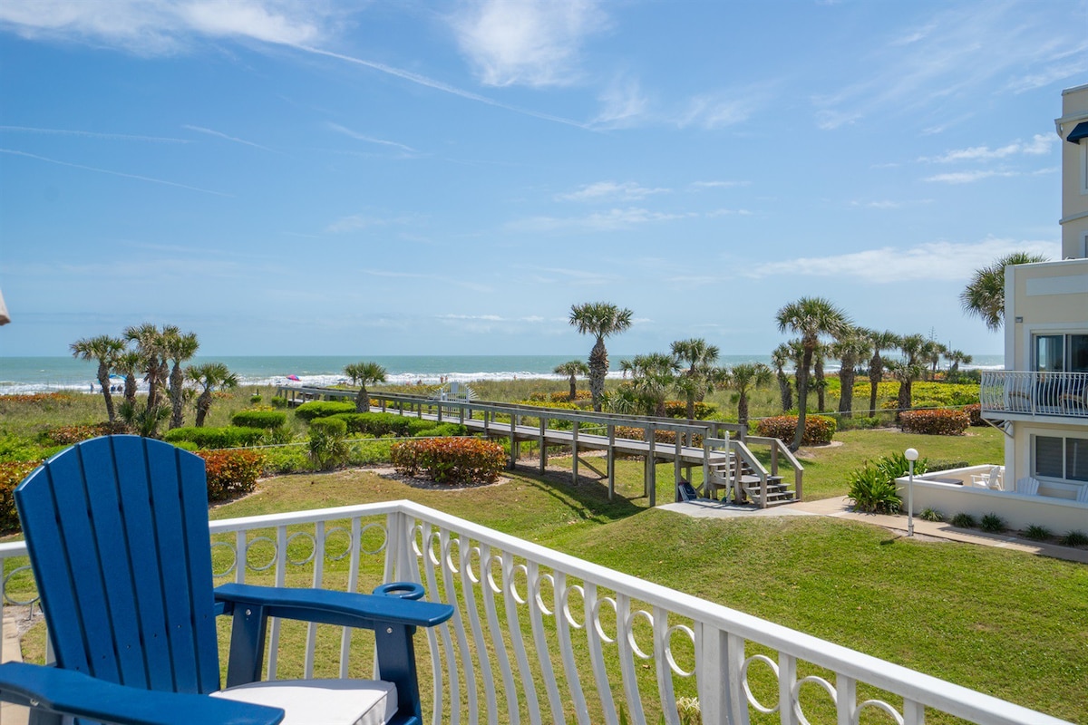 A southeast-facing balcony features a blue chair overlooking a landscaped area. The view includes a wooden walkway leading to the beach, with palm trees dotting the shoreline and the ocean visible in the background under a bright sky.