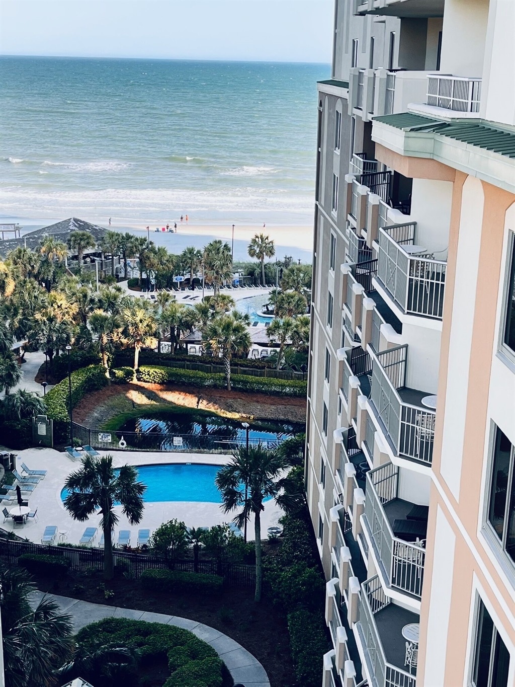 An aerial view shows a serene ocean scene with rolling waves meeting a sandy beach. Lush palm trees and a sparkling pool are visible, complementing the well-maintained greenery throughout the property. Balconies of the building are seen, providing a glimpse of the inviting outdoor spaces.