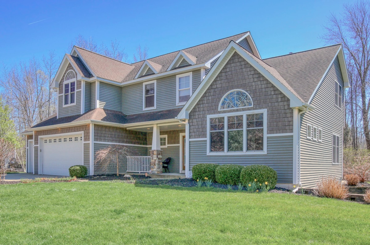 The exterior of a two-story house is featured, showcasing a combination of shingle and vinyl siding. A spacious front porch with seating is highlighted, while a manicured lawn with shrubbery surrounds the property. Tall trees are visible in the background, offering a serene setting.