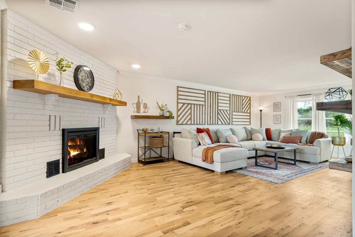 A spacious living area features light hardwood flooring and a cozy fireplace set within a white brick wall. The comfortable sectional sofa is arranged around a coffee table, with decorative accessories placed on a wooden shelf and side table. Natural light enters through large windows.
