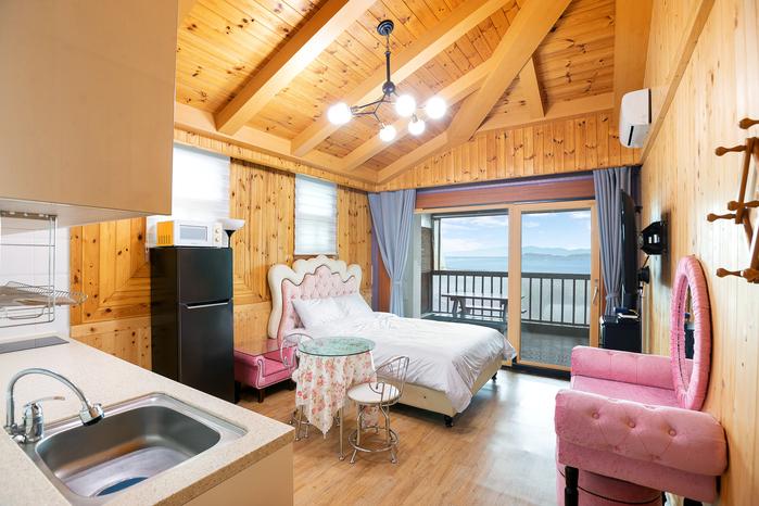 An inviting studio space features a spacious bed with a light-colored headboard, accompanied by pink furniture, including a charming chair and side table. Large windows allow natural light to fill the room, while wooden beams on the ceiling add architectural interest.