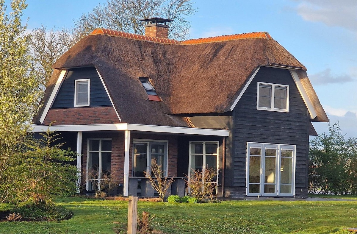 A charming two-story home features a thatched roof and dark wood exterior. Large windows allow natural light to illuminate the interior, while surrounding greenery enhances the outdoor space. The front porch provides an inviting area for relaxation amidst the well-maintained yard.