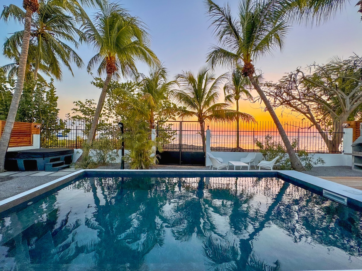 A serene pool area is framed by swaying palm trees, as the sunset casts a warm glow over the scene. The still water reflects the colorful sky, enhancing the tranquil atmosphere. A white lounge chair and modern furniture are positioned nearby, inviting relaxation.