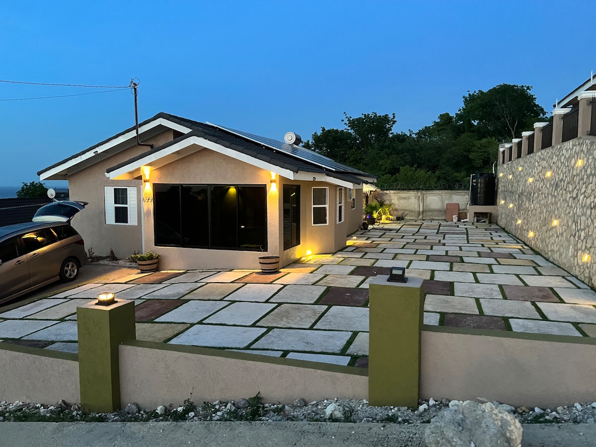 A well-lit exterior of a single-story house is presented, featuring large windows that reflect the evening sky. The driveway is paved with stone squares, bordered by green grass. A low stone wall enhances the property, while illuminated pathways suggest a welcoming ambiance.