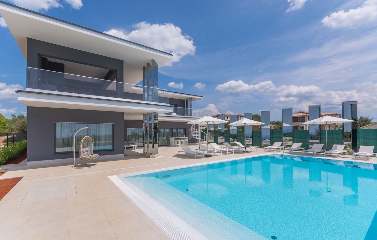 The modern villa presents a spacious outdoor area with a heated swimming pool surrounded by 10 deckchairs and three umbrellas. The clean lines of the two-story structure are complemented by expansive glass windows that invite natural light, offering views of the surrounding greenery.