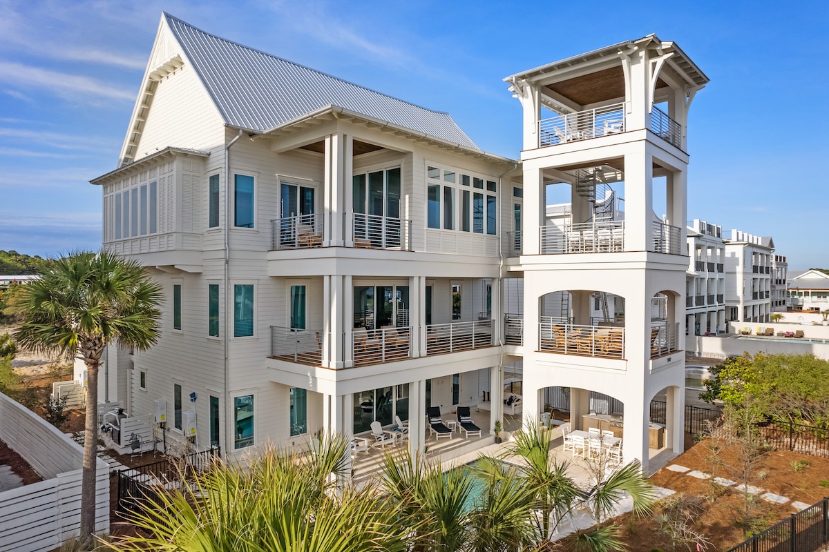 The newly constructed beach house stands with multiple levels, framed by palm trees. Balconies offer seating arrangements and views of the surrounding area. A private pool is visible in the backyard, with the structure's light-colored exterior contrasting against the blue sky.