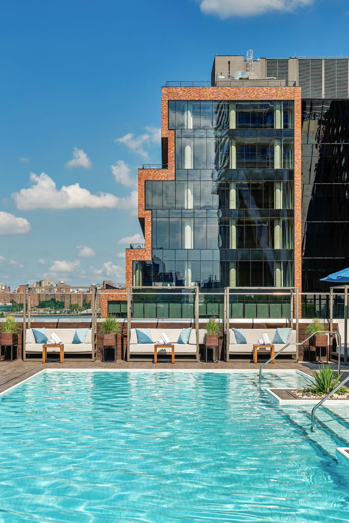 A refreshing outdoor pool is complemented by wooden decking and lounge chairs that invite relaxation. The modern building is characterized by its striking brick and glass facade, reflecting the blue sky and clouds above.