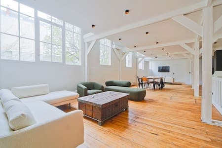 The spacious living area features large windows allowing natural light to fill the room. A combination of soft seating options, including a sectional sofa and unique green armchairs, surrounds a rustic coffee table. A dining table is set against the wall, with minimalist decor enhancing the open layout.