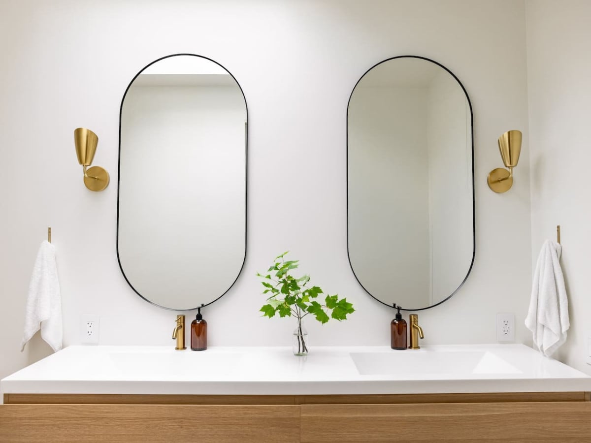 A modern bathroom features a double sink with a clean white countertop and warm wooden cabinetry. Two large, rounded mirrors are mounted above the sinks, reflecting a minimalist design. A small green plant is placed between the mirrors, and elegant wall sconces provide subtle lighting.