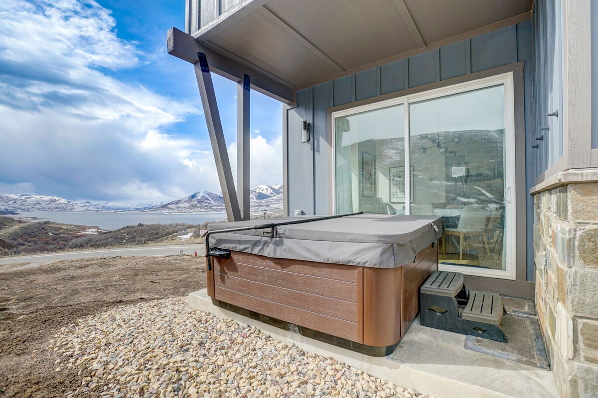 A hot tub is situated on a private patio, offering views of the surrounding landscape. The exterior features a mix of wooden and stone elements, while expansive windows allow for natural light to enter the interior space. The nearby mountains are visible in the distance.