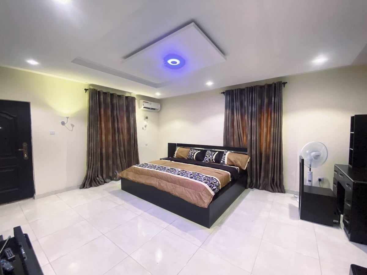 A spacious bedroom is furnished with a large bed, dressed in neutral and patterned linens. Soft lighting is provided by ceiling fixtures and curtains frame the windows. A desk and chair are situated against the wall, alongside a fan for added comfort.