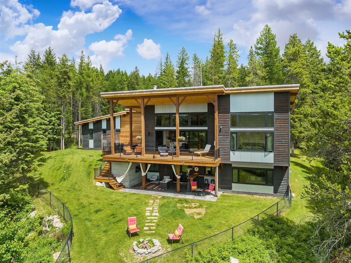 A modern home is set within a lush green landscape, featuring multiple levels and large windows. A spacious deck extends from the main structure, with outdoor seating visible. A fire pit area is framed by stone pathways, while surrounding trees enhance the serene environment.
