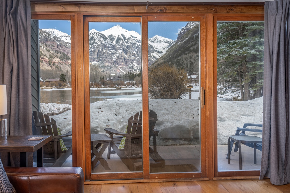 Large sliding glass doors frame a stunning view of snow-covered mountains and a serene landscape. Comfortable outdoor seating, including Adirondack chairs, is positioned on the private deck, inviting relaxation while taking in the surrounding natural beauty.
