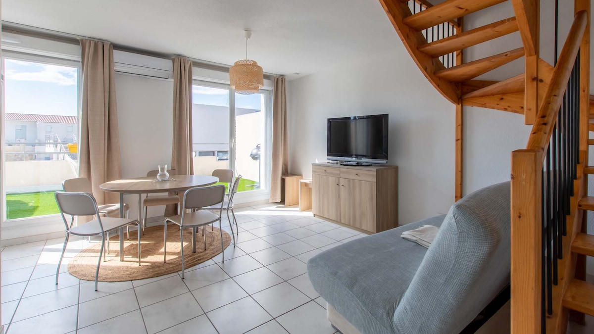 A bright living area features a light-colored sofa bed and a wooden TV stand opposite a flat-screen television. A round dining table with four chairs is situated next to large windows that allow natural light to fill the space, enhancing the airy ambiance.