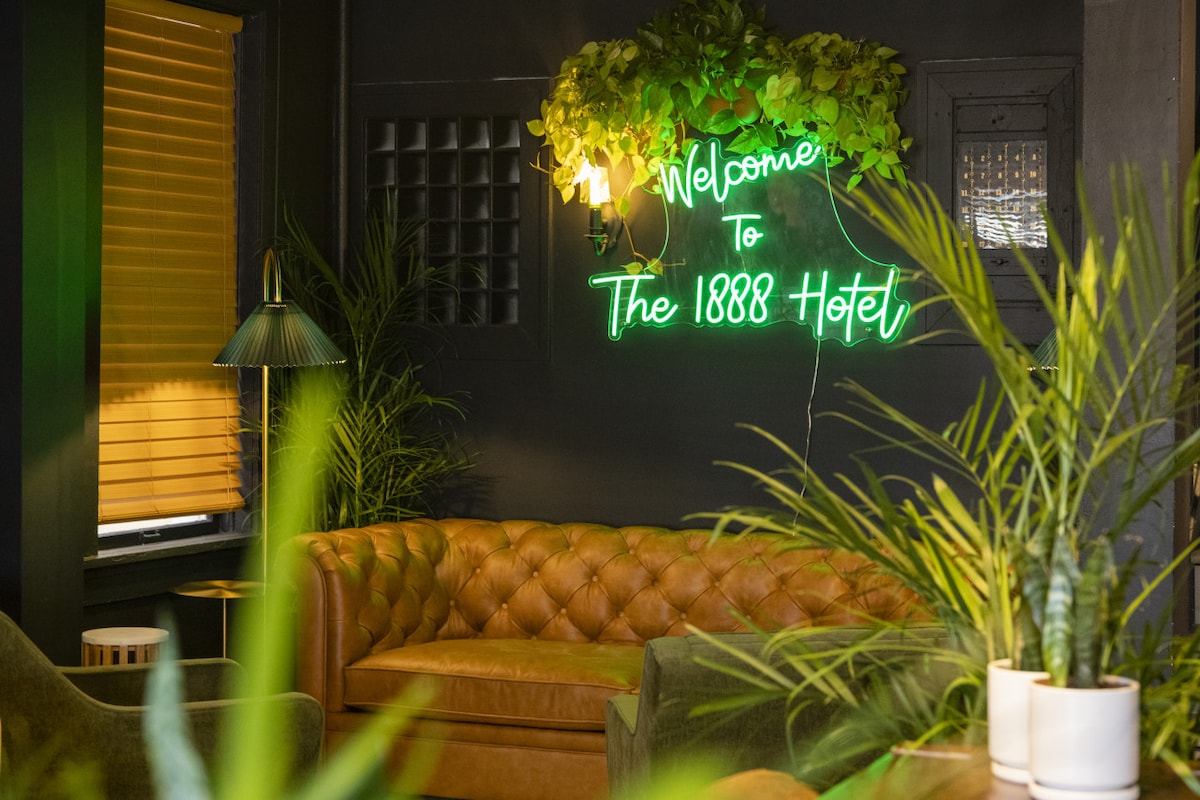 A welcoming lounge area features a brown leather couch paired with green foliage. A neon sign displays the message 'Welcome to The 1888 Hotel.' Ambient lighting is provided by a stylish lamp, creating a comfortable and inviting space.