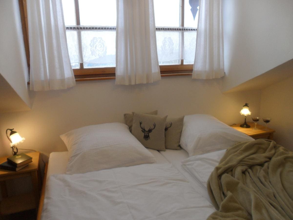 A cozy sleeping area is presented with a spacious bed featuring clean, white linens. Soft pillows are arranged against a backdrop of light curtains with a subtle pattern. Warm bedside lamps emit a gentle glow, and a plush blanket adds a touch of comfort.