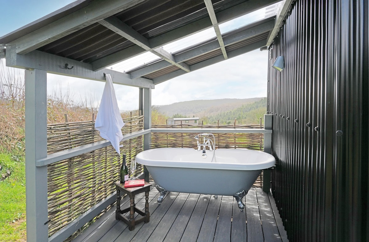 An outdoor bathtub is positioned on a wooden deck, accompanied by a small table. A white towel hangs nearby, and the space is surrounded by a natural bamboo fence. Scenic views of green hills and valleys are visible in the background.