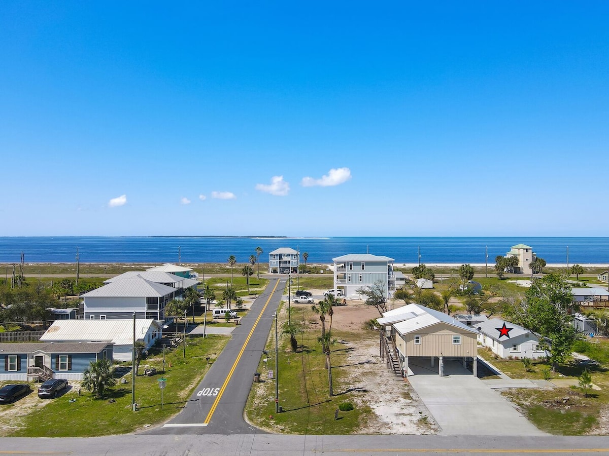 An aerial view showcases a peaceful coastal neighborhood, featuring various homes and the distant Gulf of Mexico. Palm trees dot the landscape, and a clear blue sky enhances the serene atmosphere. Streets lead towards the water, indicating proximity to the beach.